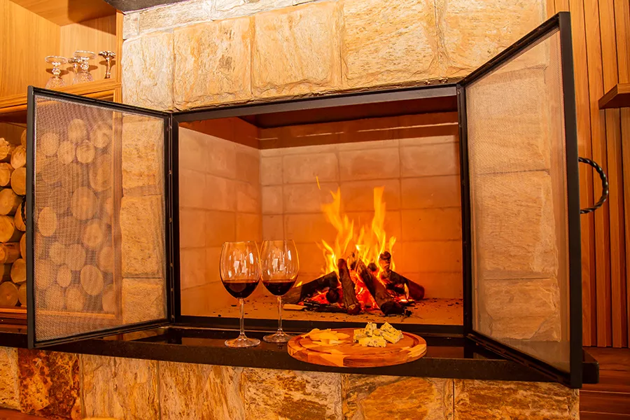 Stone fireplace with open mesh doors, a fire burning inside, two glasses of red wine and a wooden platter with cheese and crackers on the hearth.